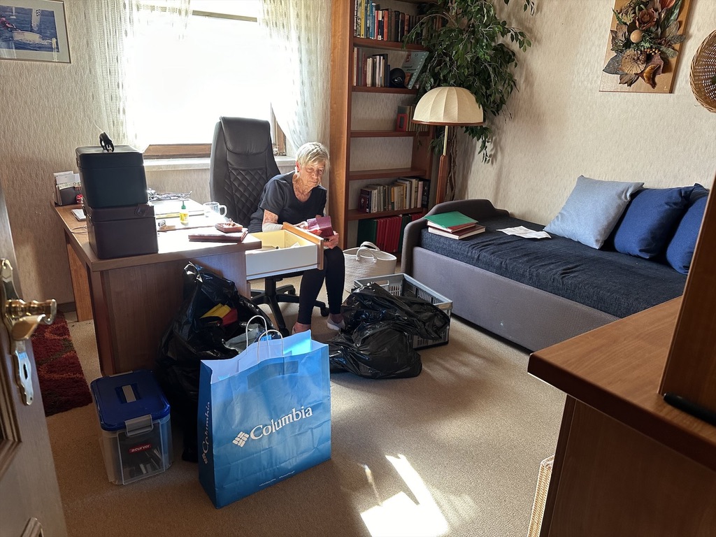 Ein Zimmer wird geräumt, Taschen und Säcke stehen herum. Eine alte Frau sortiert Unterlagen aus.